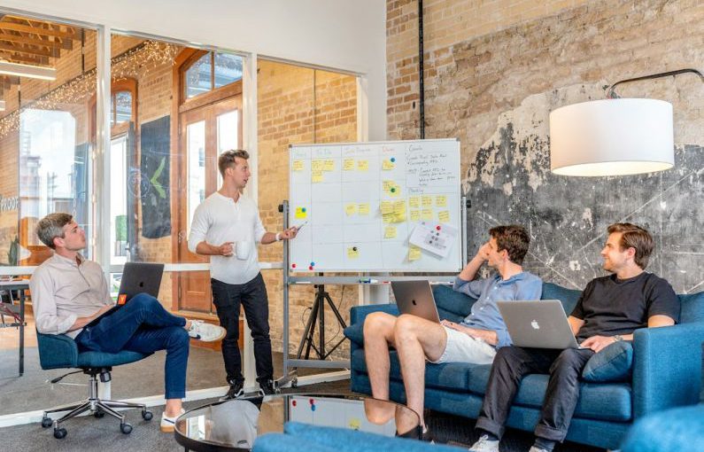 Startups - three men sitting while using laptops and watching man beside whiteboard
