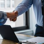 Collaborative Workspace - Crop unrecognizable coworkers in formal wear standing at table with laptop and documents while greeting each other before meeting