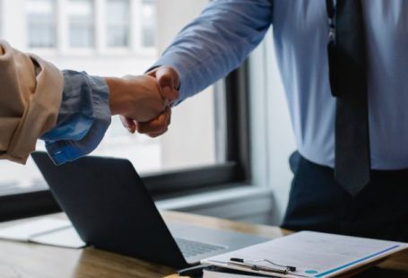 Collaborative Workspace - Crop unrecognizable coworkers in formal wear standing at table with laptop and documents while greeting each other before meeting