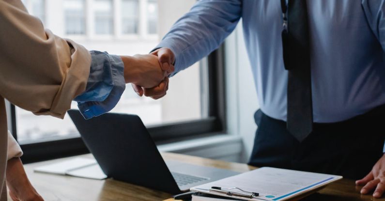 Collaborative Workspace - Crop unrecognizable coworkers in formal wear standing at table with laptop and documents while greeting each other before meeting