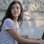 Fashion Startup - Side view of ethnic female freelancer in casual striped t shirt sitting at table on terrace and typing on netbook while working remotely in cafe and looking away