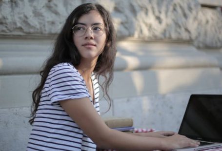 Fashion Startup - Side view of ethnic female freelancer in casual striped t shirt sitting at table on terrace and typing on netbook while working remotely in cafe and looking away