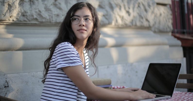 Fashion Startup - Side view of ethnic female freelancer in casual striped t shirt sitting at table on terrace and typing on netbook while working remotely in cafe and looking away