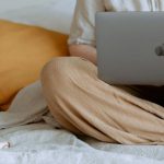 Social Startup - Crop faceless female in casual outfit sitting on bed with legs crossed holding laptop on knees with cup of coffee standing on notebook while working from home