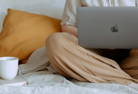 Social Startup - Crop faceless female in casual outfit sitting on bed with legs crossed holding laptop on knees with cup of coffee standing on notebook while working from home