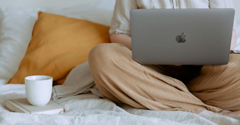 Social Startup - Crop faceless female in casual outfit sitting on bed with legs crossed holding laptop on knees with cup of coffee standing on notebook while working from home