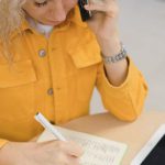 Career Fair - From above of female freelancer taking notes in notebook while sitting at table and using smartphone for making call