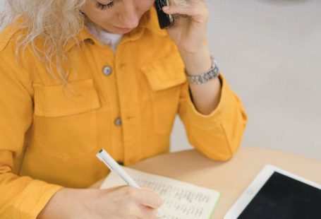 Career Fair - From above of female freelancer taking notes in notebook while sitting at table and using smartphone for making call