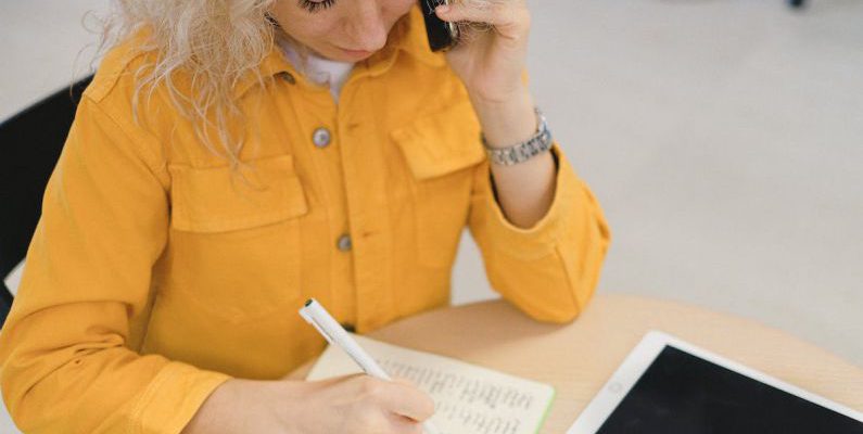 Career Fair - From above of female freelancer taking notes in notebook while sitting at table and using smartphone for making call