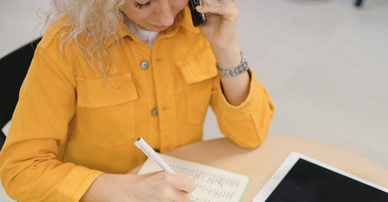 Career Fair - From above of female freelancer taking notes in notebook while sitting at table and using smartphone for making call